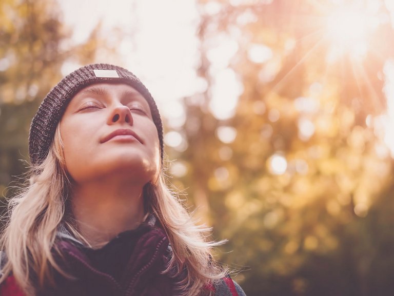 Frau im Wald neigt mit geschlossenen Augen und einem leichten Lächeln den Kopf nach hinten und wirkt entspannt