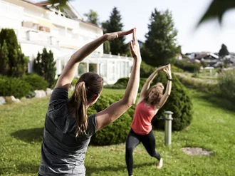 zwei Frauen die im Garten Yoga machen, im Hintergrund das Gebäude